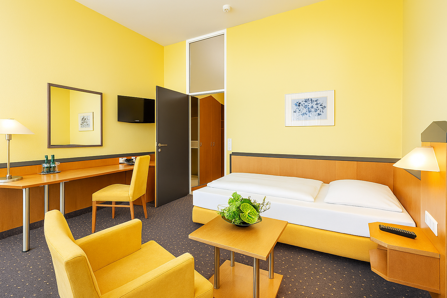 Hotelzimmer mit gelben Wänden und halbhohen Holzakzenten, gelben Stühlen und Bett. Im Raum stehen ein großer Schreibtisch und eine Sitzecke.