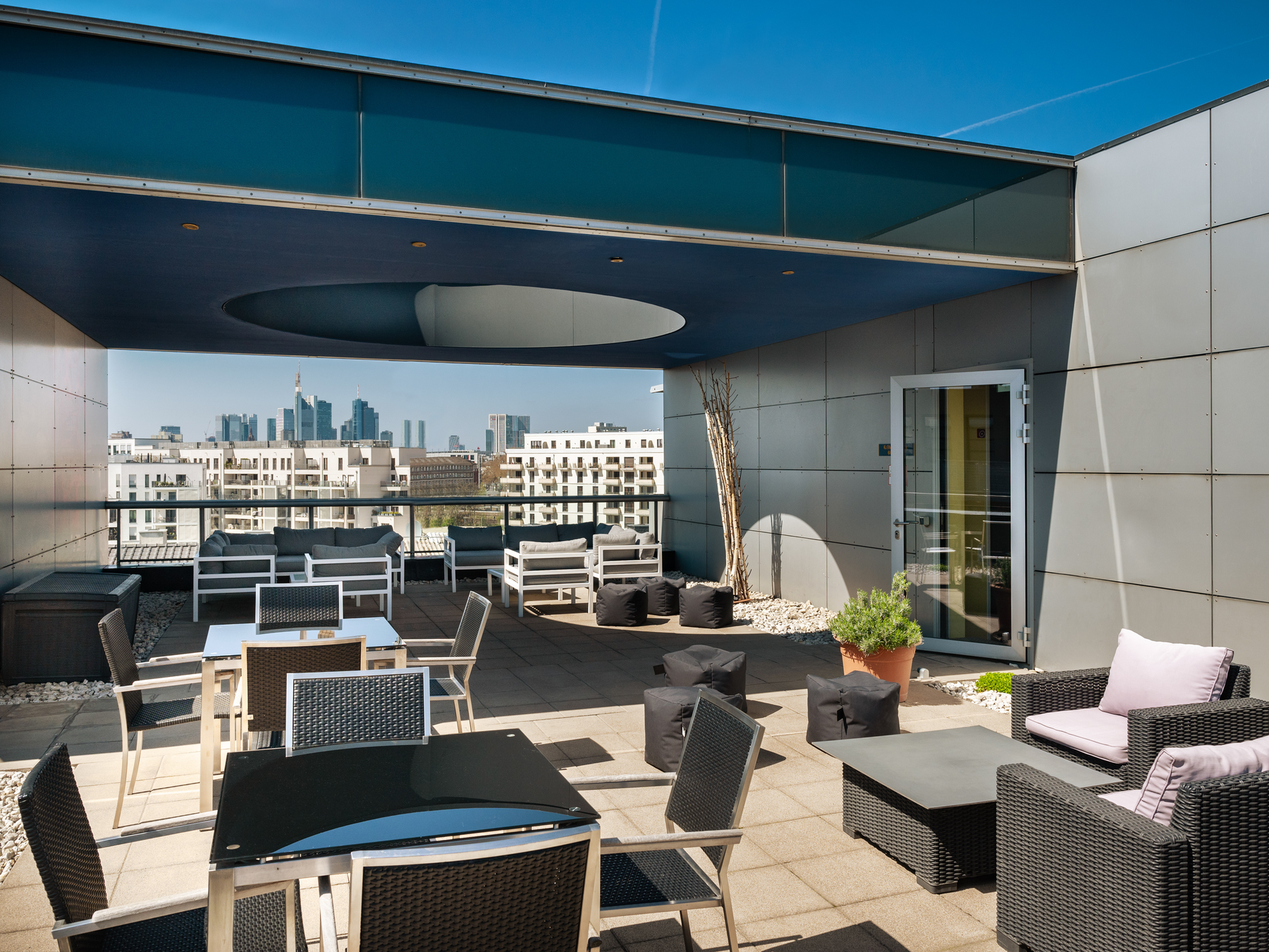 Viele Gäste auf der stilvoll möblierten, modernen Dachterrasse im warmen Licht des Abendrots. In der Decke der Terrasse ist ein kreisrunder Ausschnitt, durch den ein offener, farbenprächtiger Abendhimmel zu sehen ist.