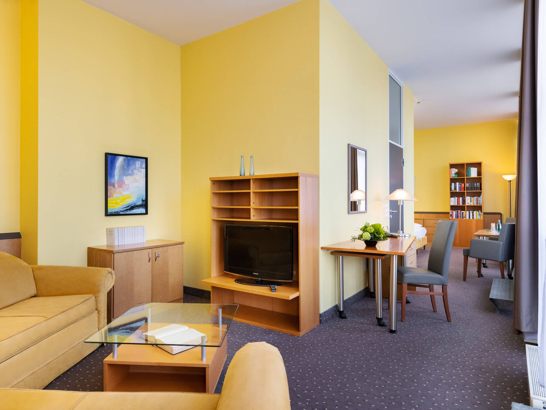 Wohnzimmer der Suite mit gelbem Sofa, Glastisch und gelben Wänden. Ein Holzregal steht im Raum, und im Durchgang zum Schlafzimmer steht ein Tisch mit Spiegel. Bodentiefe Fensterfront zur Terrasse lässt viel Licht herein.