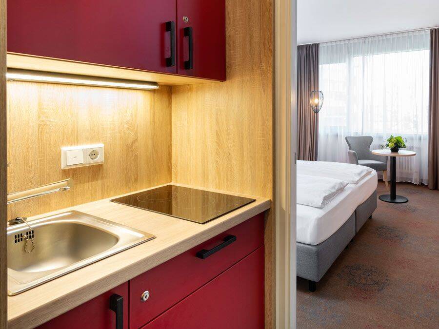 Brandneue Kitchenette mit modernen roten Fronten, hölzernen Details und Ceranfeld. Im Hintergrund erkennt man das gemütliche Hotelzimmer mit Doppelbett und Sitzecke am Fenster