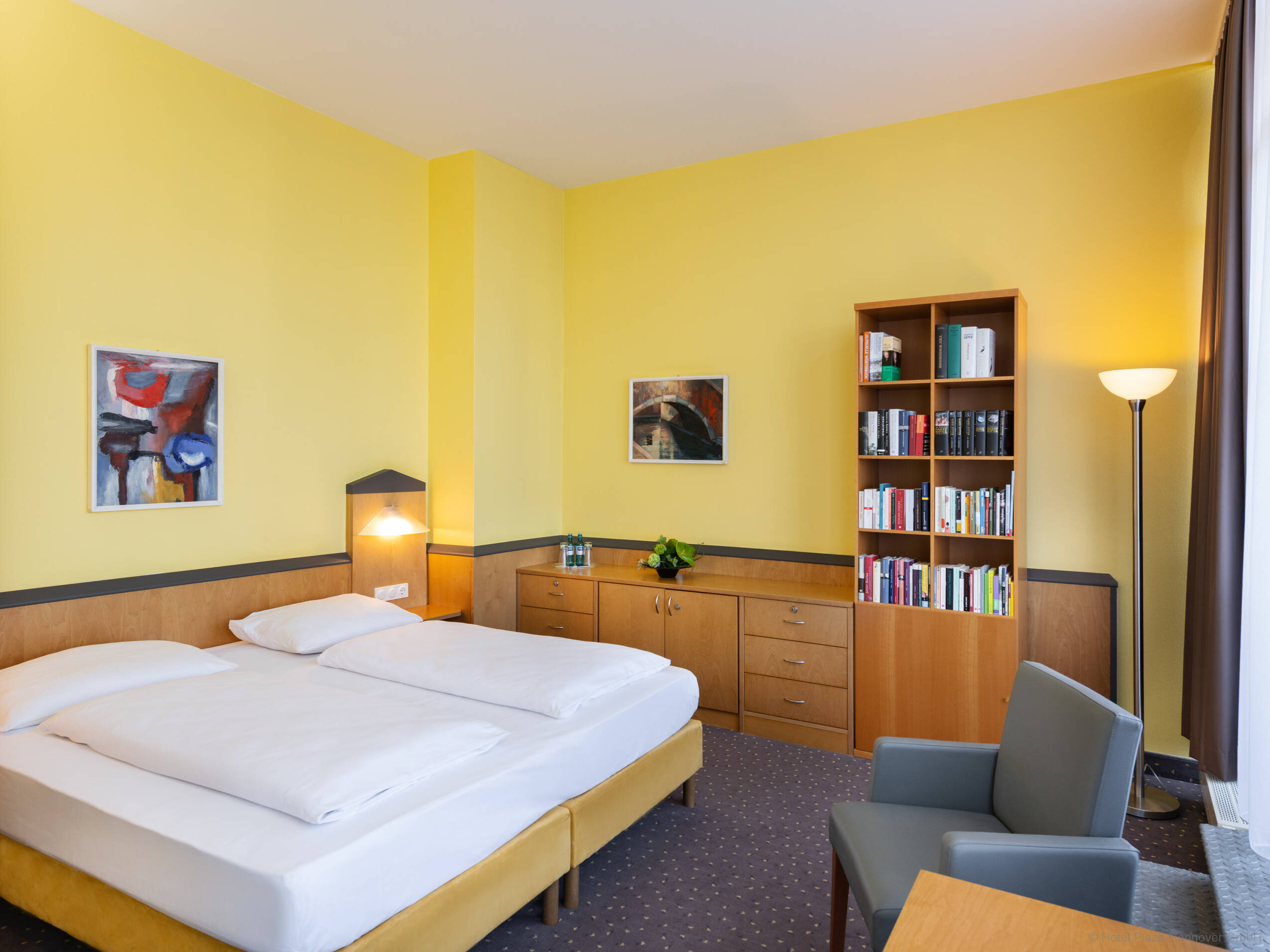 Schlafzimmer der Suite mit bodentiefer Fensterfront zur Terrasse, einem Doppelbett und gelben Wänden. Die Wände sind halbhoch mit Holz verkleidet. Neben dem Bett stehen ein Einbauschrank und ein Bücherregal mit einem Sessel.
