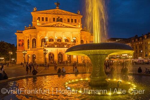 Alte Oper am Abend mit einem Brunnen und sprudelnder Fontäne im Vordergrund, in dessen Wasser sich die Lichter spiegeln. Im Hintergrund sind Passanten und das beleuchtete Operngebäude vor einem tiefblauen Nachthimmel zu sehen.