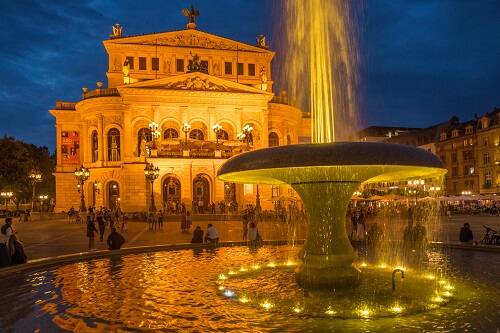 Alte Oper am Abend mit einem Brunnen und sprudelnder Fontäne im Vordergrund, in dessen Wasser sich die Lichter spiegeln. Im Hintergrund sind Passanten und das beleuchtete Operngebäude vor einem tiefblauen Nachthimmel zu sehen.