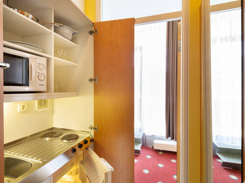 Ein Gast steht vorn der Kitchenette im Hotelzimmer und schaltet den Herd an. Der große Kühlschrank steht offen.