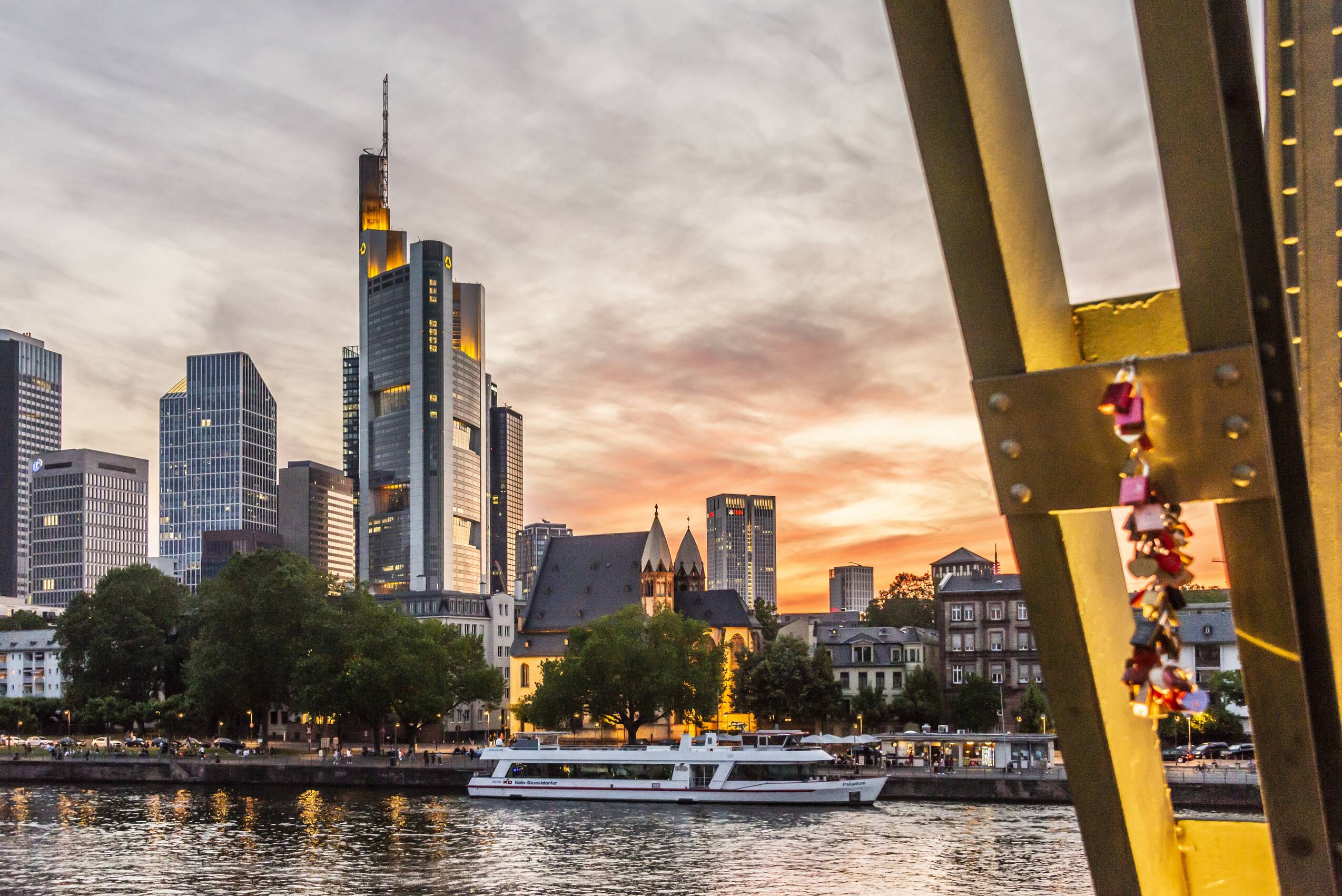 Blick auf Frankfurt vom Eisernen Steg aus. Im Vordergrund hängen einige Liebesschlösser an der Brücke, die über den Main führt. Am Ufer liegt ein Touristenschiff, und im Hintergrund ist die Skyline Frankfurts vor einem dramatischen Abendhimmel zu sehen.